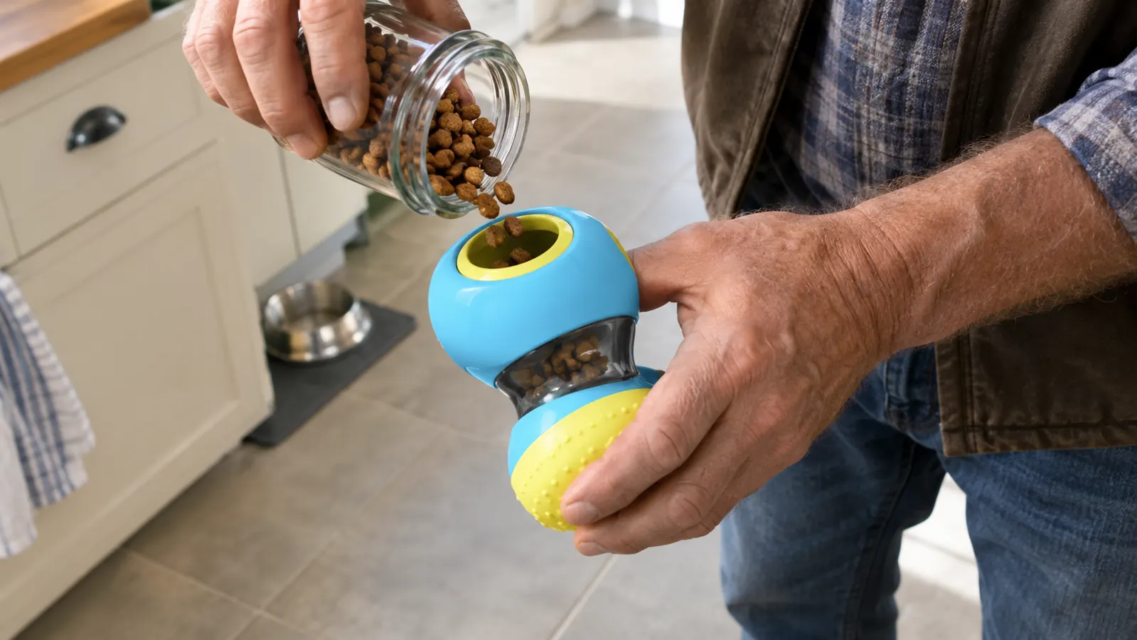 A hand sprinkling a small amount of kibble into the opening of a treat-dispensing toy on a kitchen floor